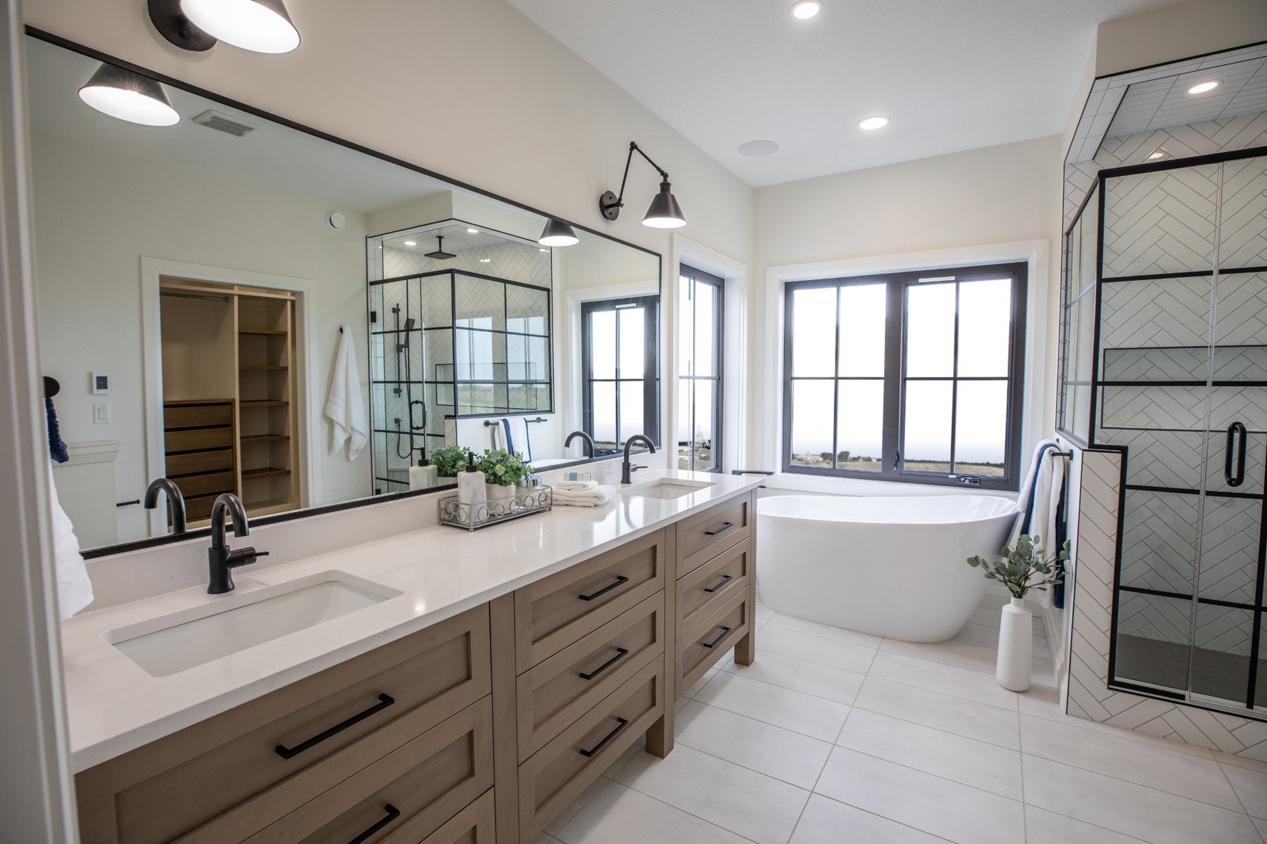 Bright primary ensuite with a light wood double vanity, freestanding tub, black-framed shower and expansive windows.