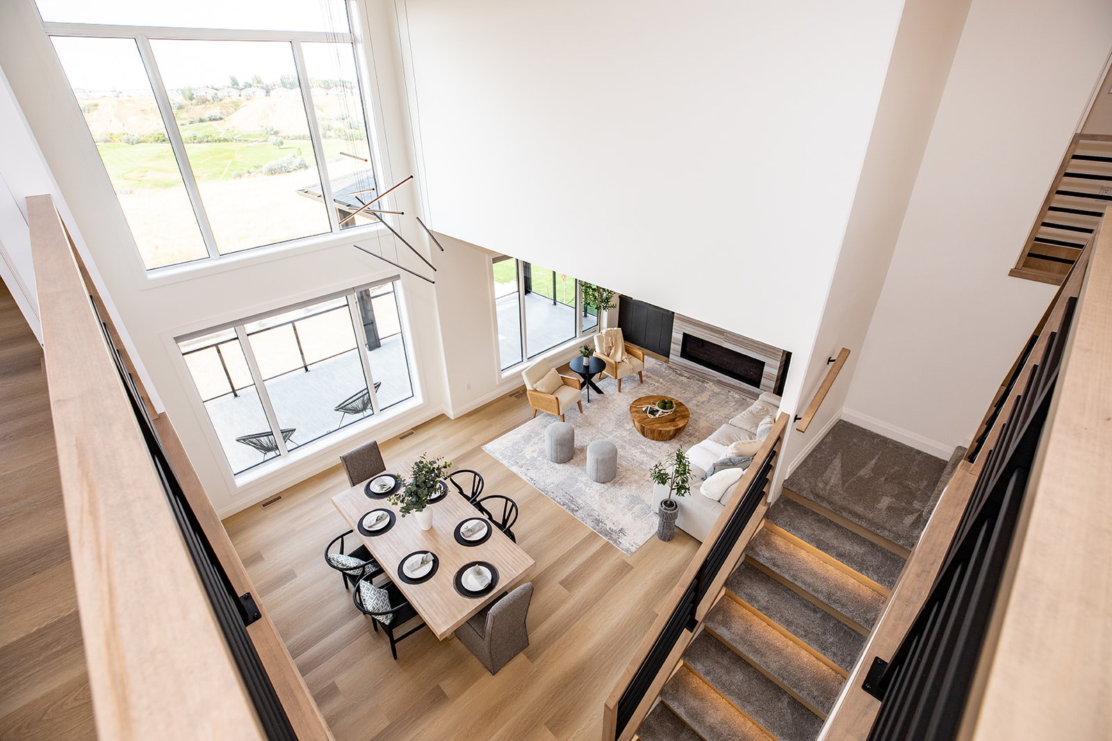 Upper-level overlook into a double-height great room and dining area with expansive windows, a fireplace and illuminated stair detailing.