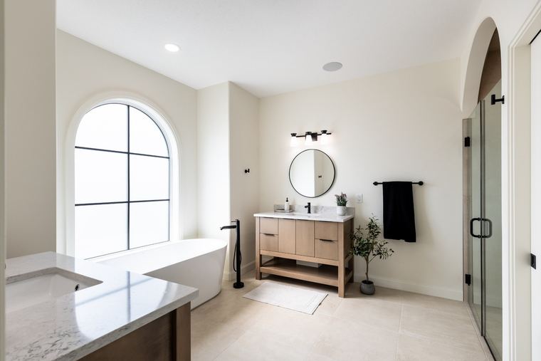 Bright primary ensuite with a freestanding tub, arched black-grid window, light wood vanity and glass shower.