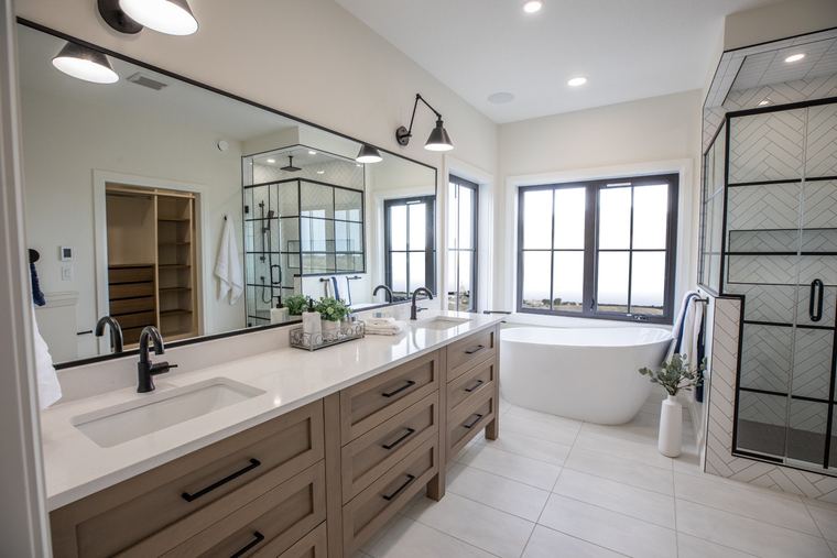 Bright primary ensuite with a light wood double vanity, freestanding tub, black-framed shower and expansive windows.